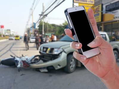 Hand holding up a cellphone with a motorcycle vs. car crash in the background.