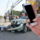 Hand holding up a cellphone with a motorcycle vs. car crash in the background.