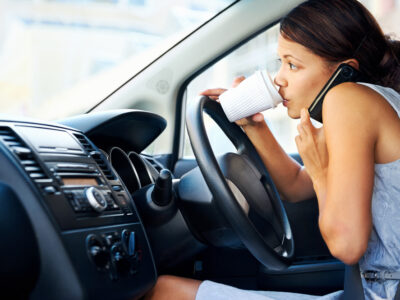 Woman in driver's seat of car hunched over the wheel drinking coffee from a paper cup and holding cellphone.